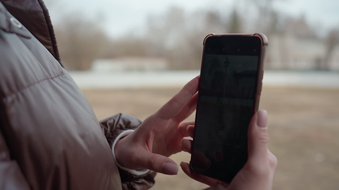 여자가 야외에서 녹화된 휴대폰 동영상을 보고 있어요. 코트와 겨울옷을 껴입고 있으며, 엄지손가락으로 화면을 스크롤하여 재생하고 있어요. 배경에는 앙상한 나무와 흐릿한 하늘이 있어 조용하고 사색적인 분위기예요.