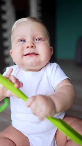 cute funny baby boy in white bodysuit sits on the floor. blue-eyed child takes kitchen spatula and smiles happily. vertical video.
