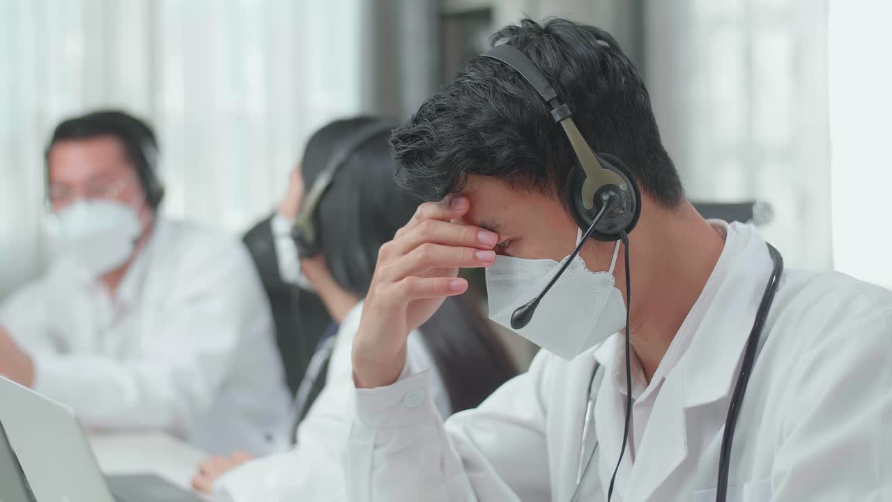 Close Up Of An Asian Man Doctor Wearing Headsets And Masks Working As Call Center Agent Is Tired Because His Colleagues Are Talking During Working At The Office