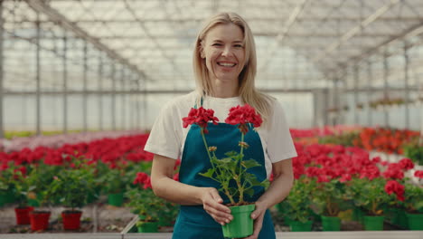 a smiling woman holding a potted red flower in a greenhouse