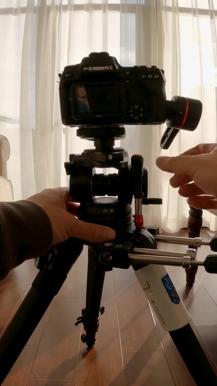 Video vertical: Manos ajustando la abrazadera de un trípode DSLR en casa para asegurar el equipo, con una camiseta marrón.