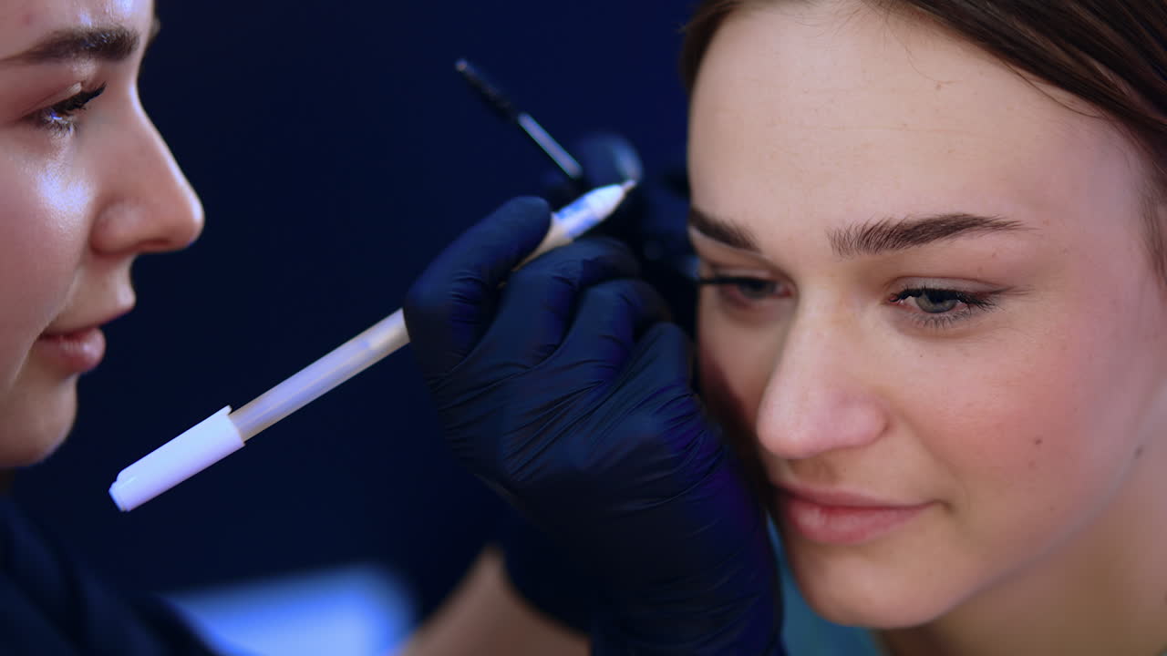 Young beautiful girl in a beauty salon. Beautician draws white lines around client's eyebrows to mark their limits for future procedure. Close up.