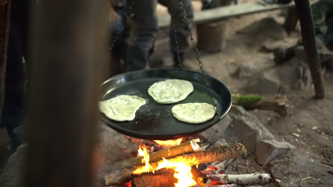 Piadine che friggono in una padella sospesa sopra un falò acceso. Scena di cucina all'aperto con fiamme, braci e un'atmosfera rustica da campeggio nella foresta.