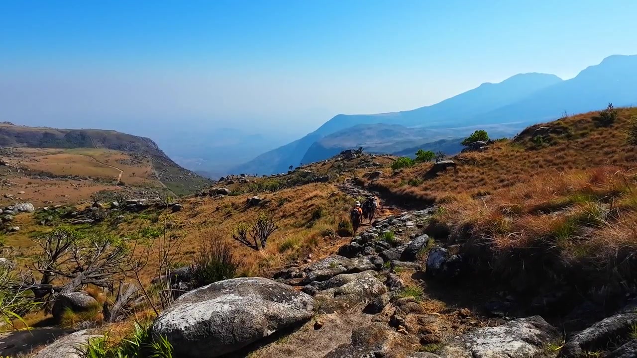 Due uomini camminano lungo un sentiero roccioso tra praterie dorate sul Monte Mulanje, Malawi, con creste montuose in lontananza sotto un cielo azzurro