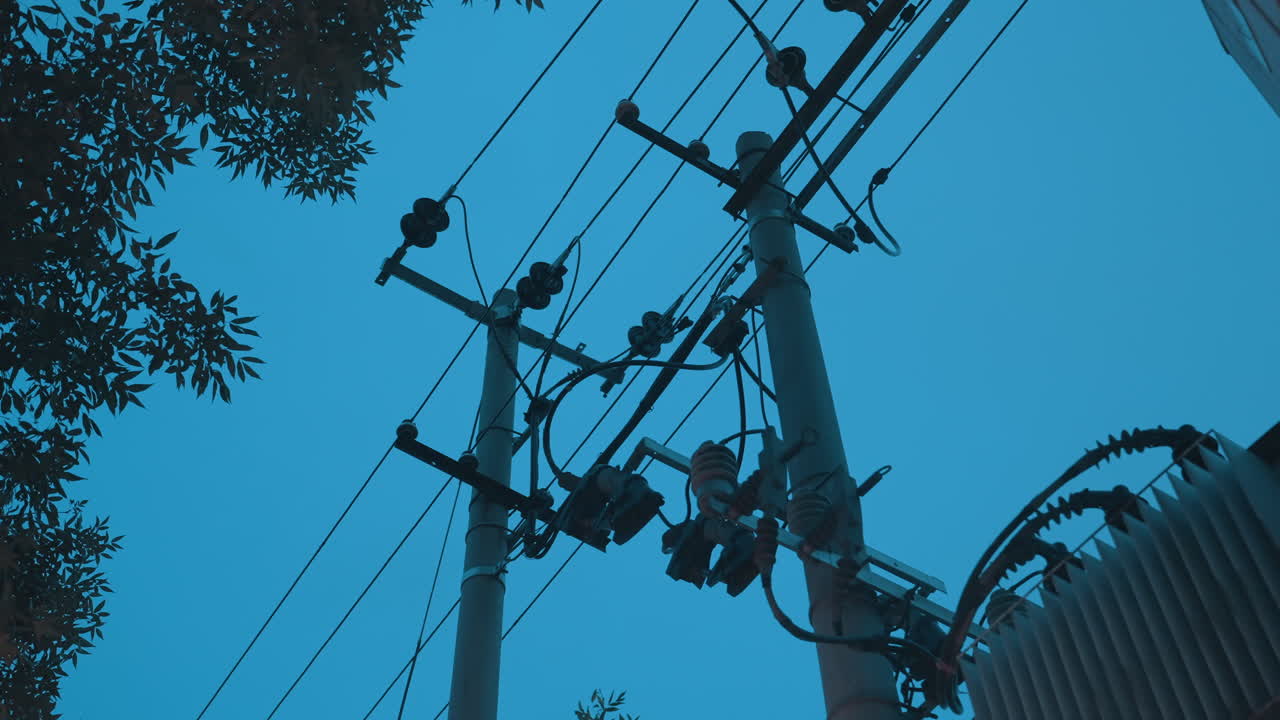 Stožár elektrického vedení, kabely transformátoru, modrá obloha, vysoce kontrastní městská silueta ukazující distribuční transformátor, izolátorové řetězce, příčník a elektrické vedení proplétající se korunami stromů, filmová nálada.