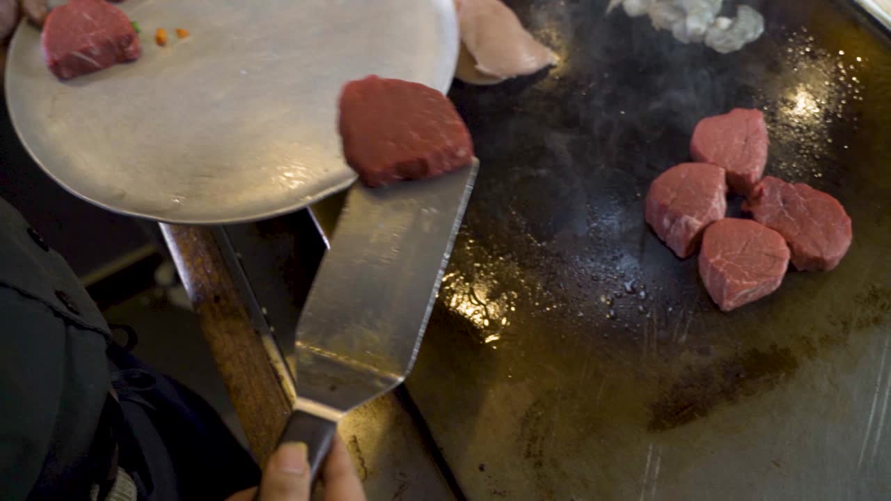 Chef moving steak from tray to hot flat grill using steel spatula, slow motion