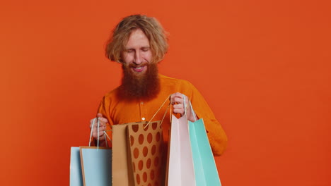 hombre caucásico mostrando bolsas de compras anunciando descuentos sonriendo pareciendo asombrado por los bajos precios