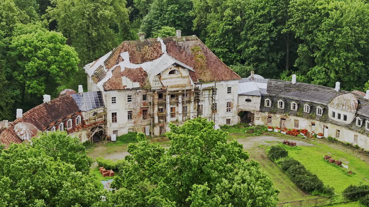 Aerial drone glides slowly past decaying Kaucminde manor framed by green trees
