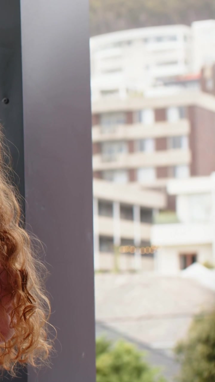 Video verticale: Panoramica su donna dai capelli rossi con top rosa che guarda appartamenti e alberi da una finestra di metallo, spazio per il testo.