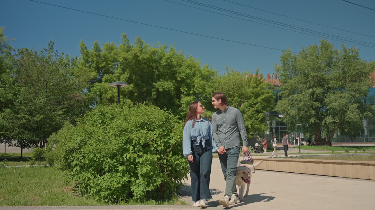 pareja blanca paseando con un labrador en un parque soleado, árboles exuberantes y camino pavimentado, ambiente relajado de fin de semana, ropa informal, banco y vistas al estanque, vegetación cálida y ambiente de ocio