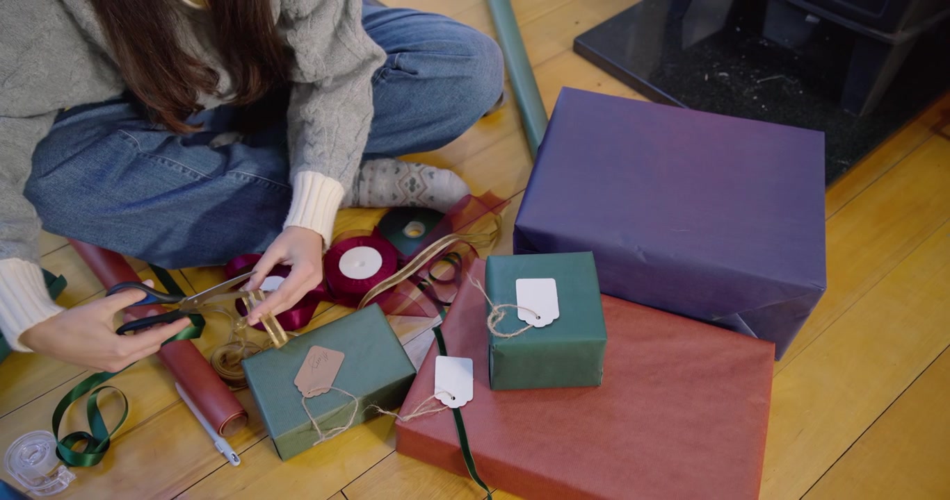 Donna che indossa un maglione grigio, che impacchetta scatole su un pavimento di legno, attaccando un'etichetta kraft e preparando regali.