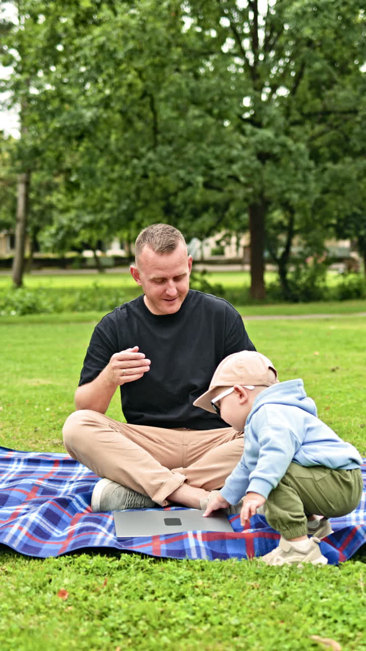 Far og lille søn sidder på græsset i haven og taler, mens de bruger en bærbar computer til at tage noter om barndomsdrømme. Ung far og hans søn på tur i parken