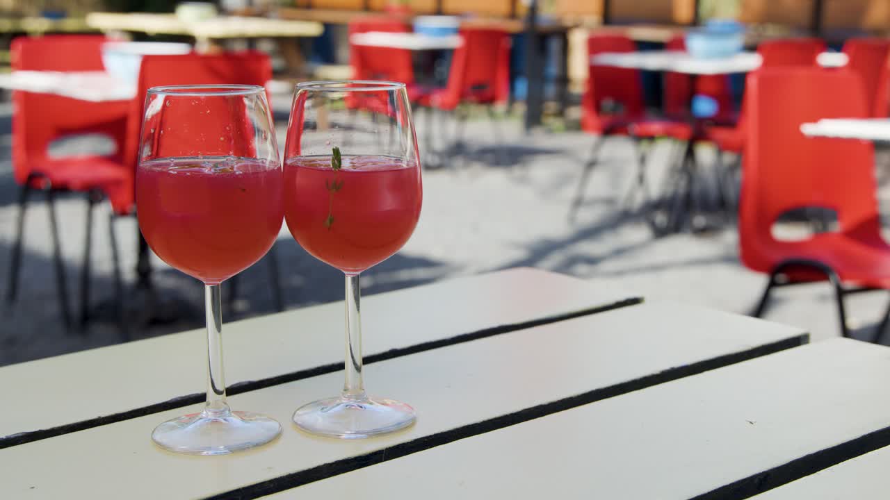 Two hands clink glasses filled with red summer cocktails at an outdoor table, surrounded by red chairs in bright natural daylight