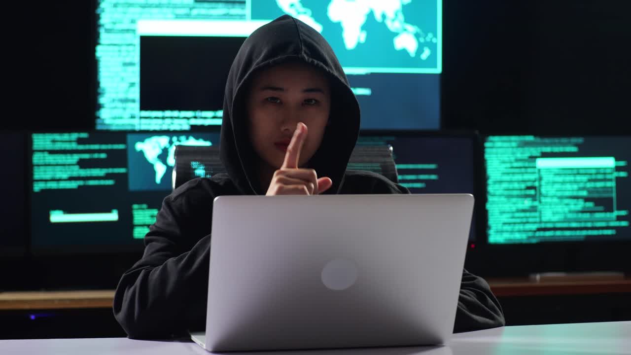 hacker asiática con capucha mostrando el gesto de "shh" con el dedo índice cerca de la boca mientras trabaja en una computadora portátil con mapas y datos en pantallas de múltiples monitores en una habitación de oficina oscura