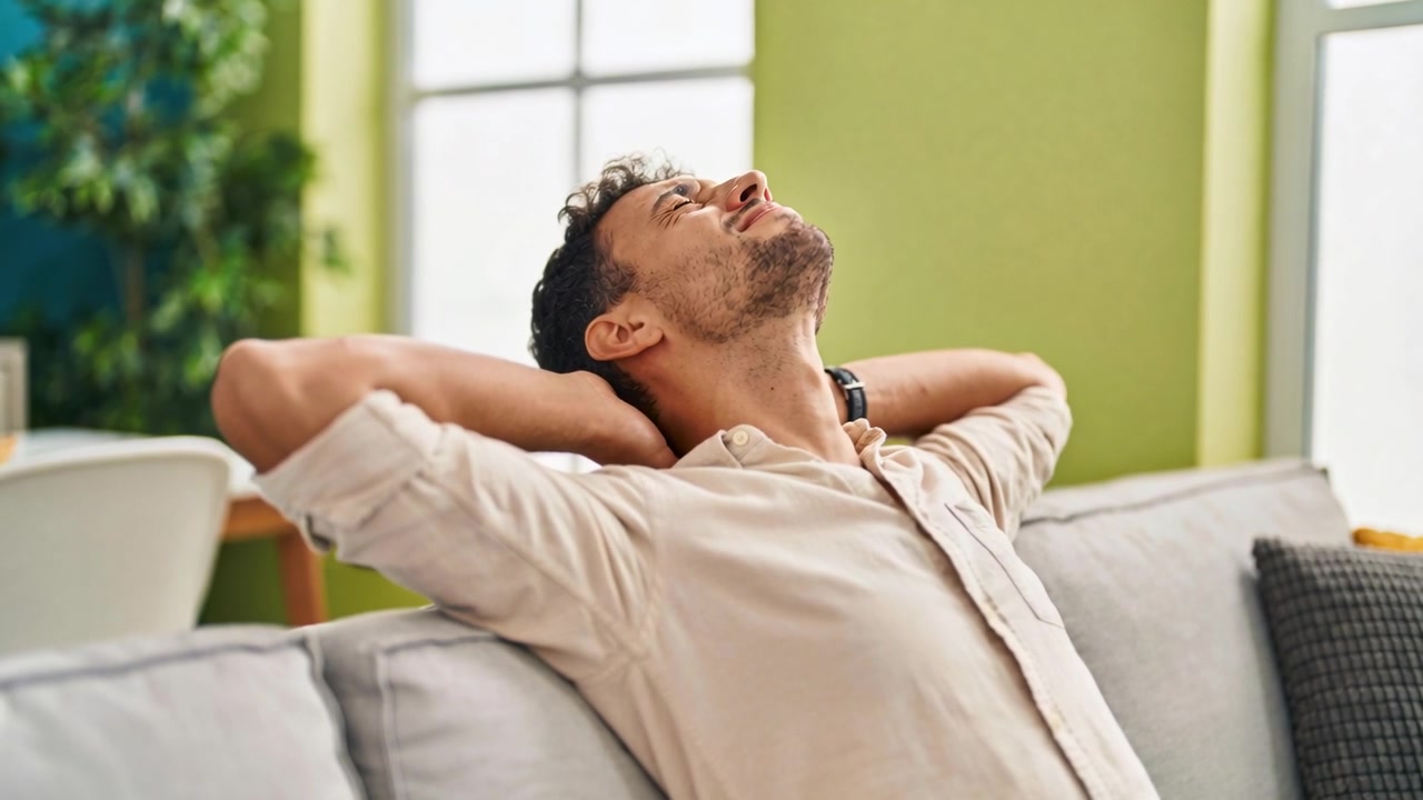 Sentado en el sofá de casa, un hombre con las mangas remangadas y un reloj inteligente, descansando y echando la cabeza hacia atrás.