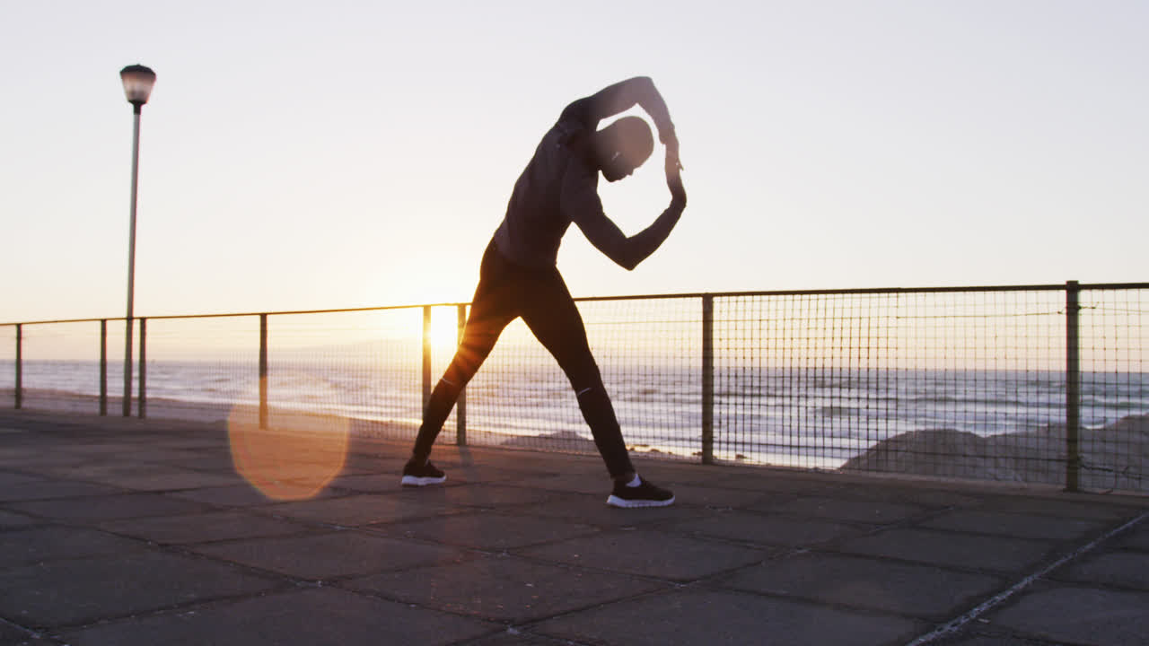 Focused african american man exercising outdoors, stretching by seaside at sunset