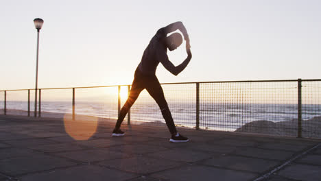 focused african american man exercising outdoors, stretching by seaside at sunset