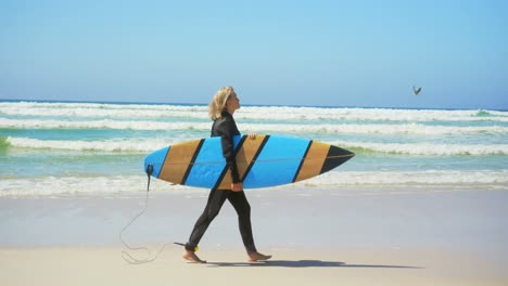 Vista-Lateral-De-Una-Surfista-Caucásica-Senior-Activa-Caminando-En-La-Playa-Bajo-El-Sol-4k