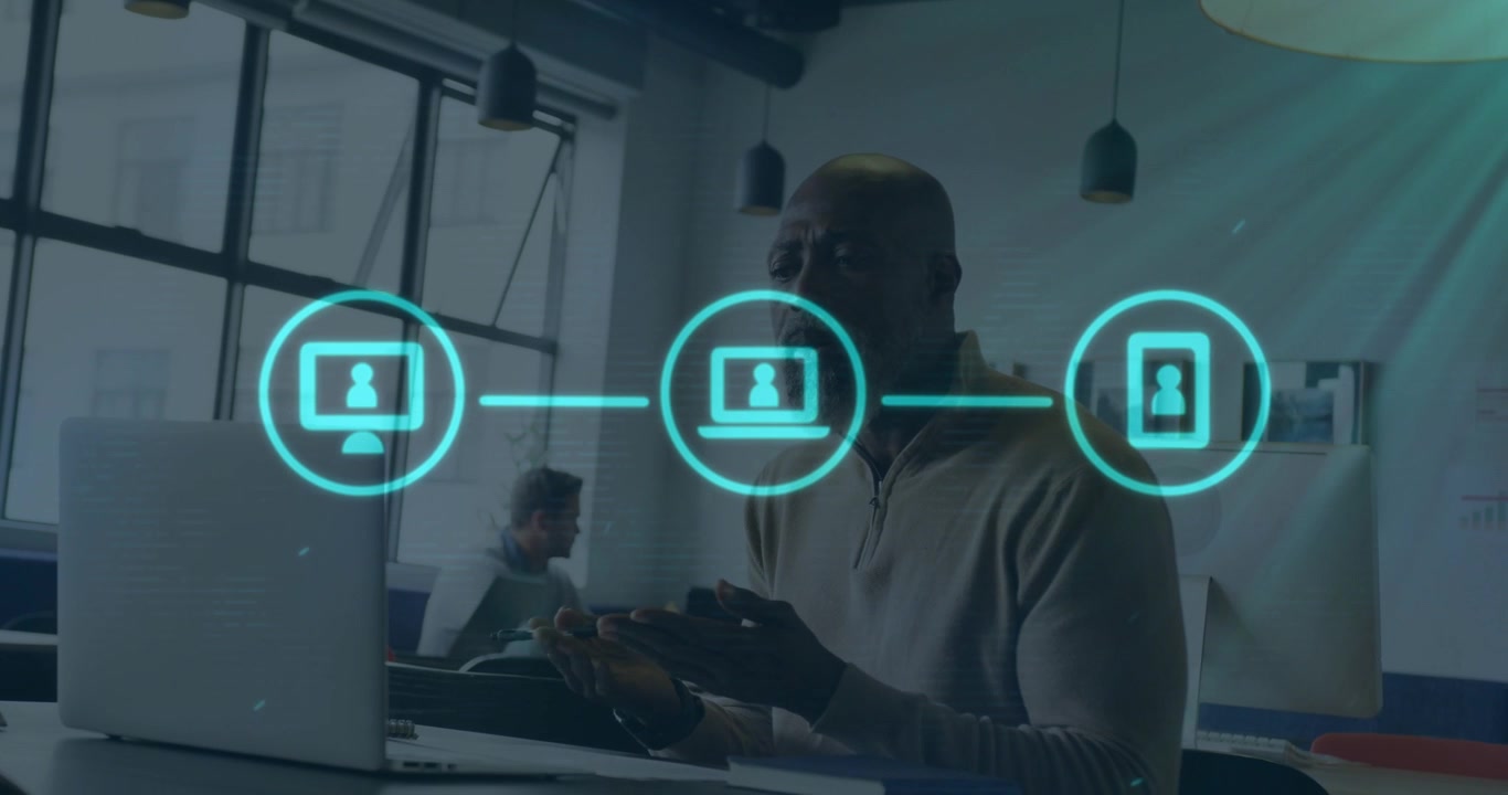 Mature male gesturing at laptop with phone causing holo icons animating across then fading for tech