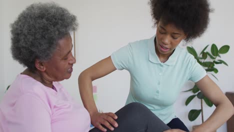 african american female physiotherapist wearing face mask helping senior female patient exercise