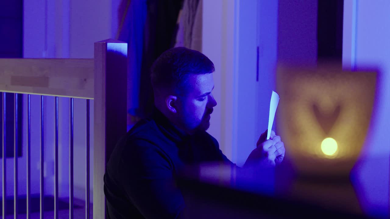 Scena cinematografica intensa di un uomo seduto da solo che legge una lettera in una luce ambientale blu, con il bagliore caldo di una candela sfocato in primo piano: concept per una rottura sentimentale