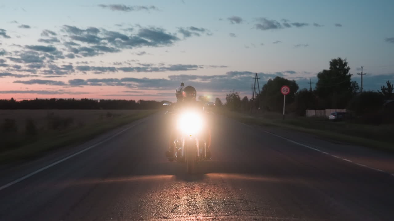 Night racer speeding on open rural road under fading sunset sky, motorcycle headlight shining brightly as powerlines, trees, and kilometer signpost frame countryside backdrop