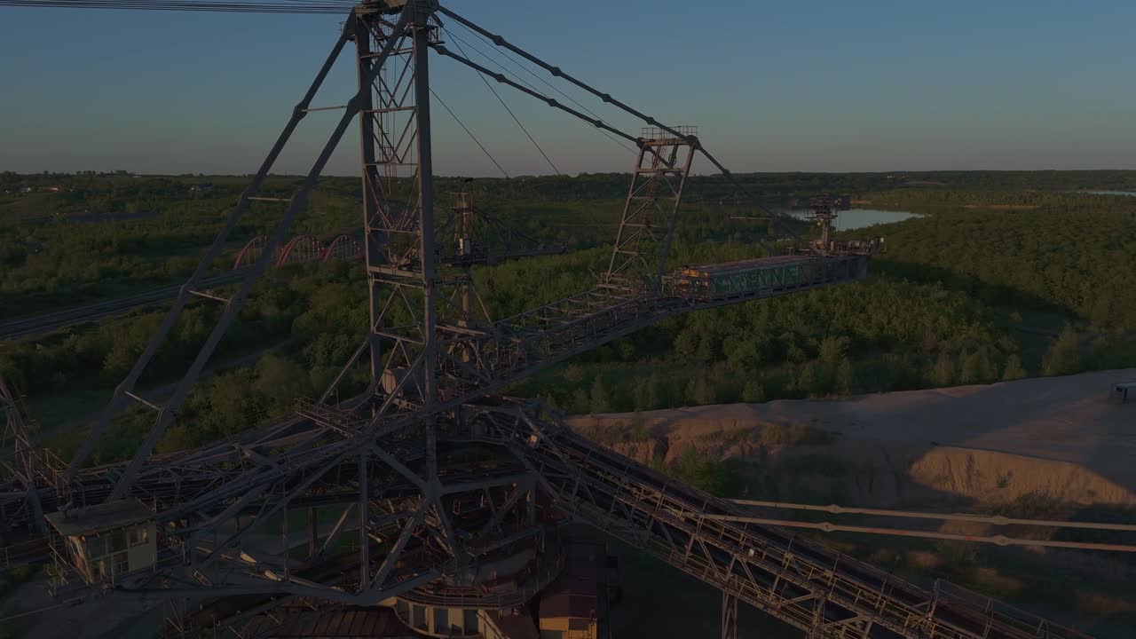Aerial sunset view of a massive industrial mining relic surrounded by forest near Leipzig, Germany. The metal colossus stands silent beside a lake, evoking the area’s mining past and transformation.