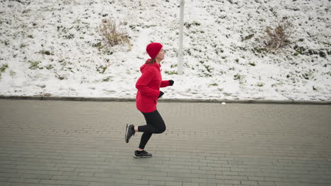 side view of coach jogging outdoors on interlocked pavement next to snowy hill during winter, in red hoodie and black leggings, serene environment featuring pole and natural snowy hillside