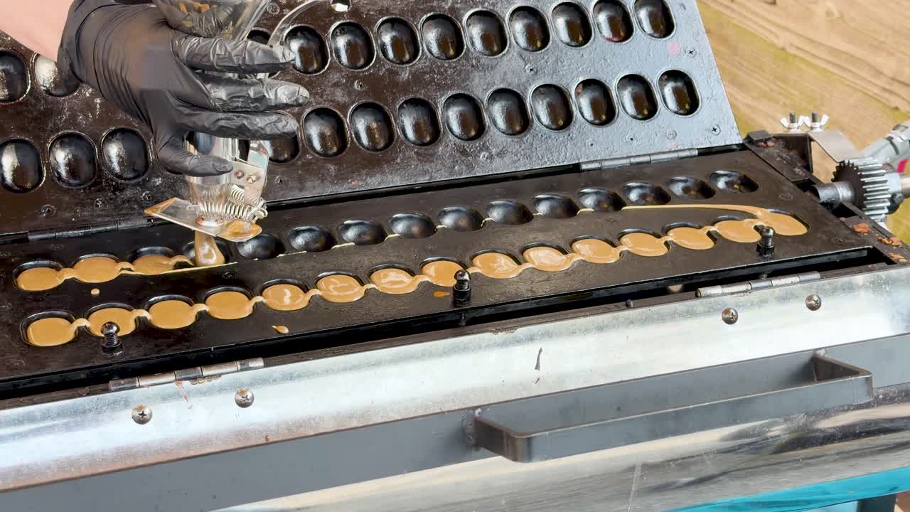 Seorang penjual makanan kaki lima bersarung tangan hitam menggunakan dispenser untuk menuangkan adonan ke atas wajan datar.