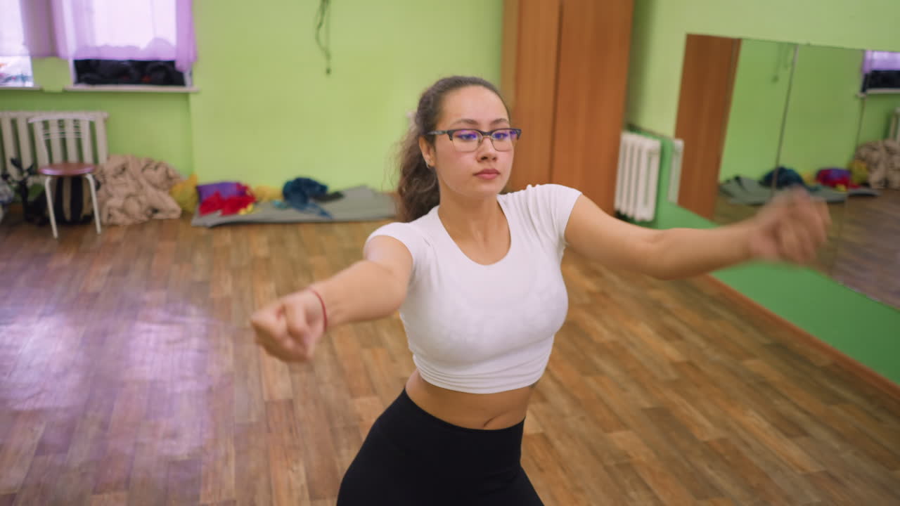 Woman in white top and eyeglasses pushing out chest during indoor workout on wooden floor gym, maintaining focus and strength while practicing fitness routine in mirrored room with energetic movement