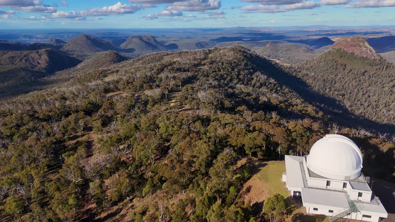 Aerial drone footage glides over a white observatory dome atop forested mountains in clear daylight, revealing sweeping views of rugged Australian wilderness