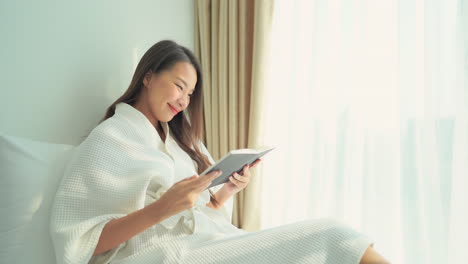 A-young-attractive-Asain-woman-sitting-up-in-her-resort-bed-turns-the-page-of-the-book-she-is-reading