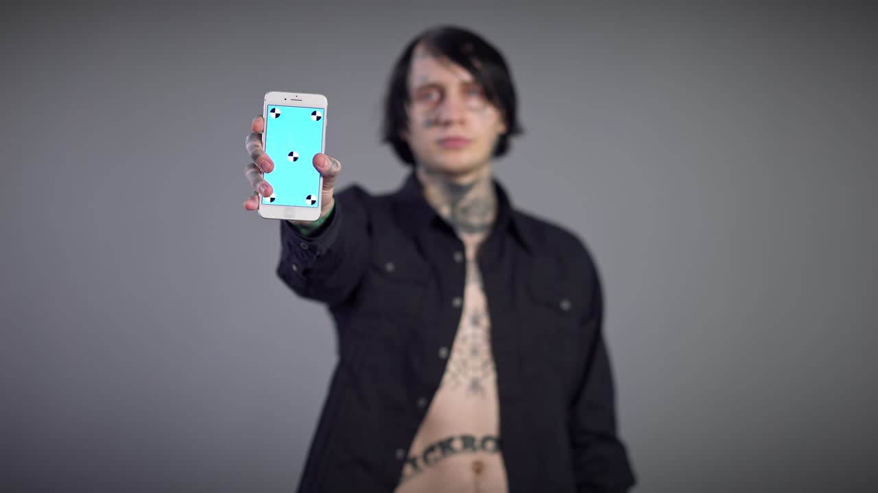 Smartphone in primo piano con schermo blu e giovane uomo caucasico tatuato sfocato sullo sfondo. Dispositivo mockup template nelle mani maschili con un ragazzo sicuro di sé in posa su sfondo grigio