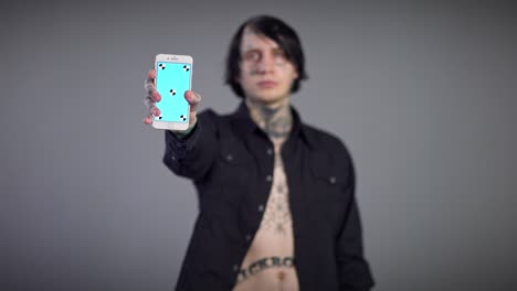 close-up smartphone with blue screen and blurred caucasian tattooed young man at background. template mockup device in male hands with confident guy posing at grey background.