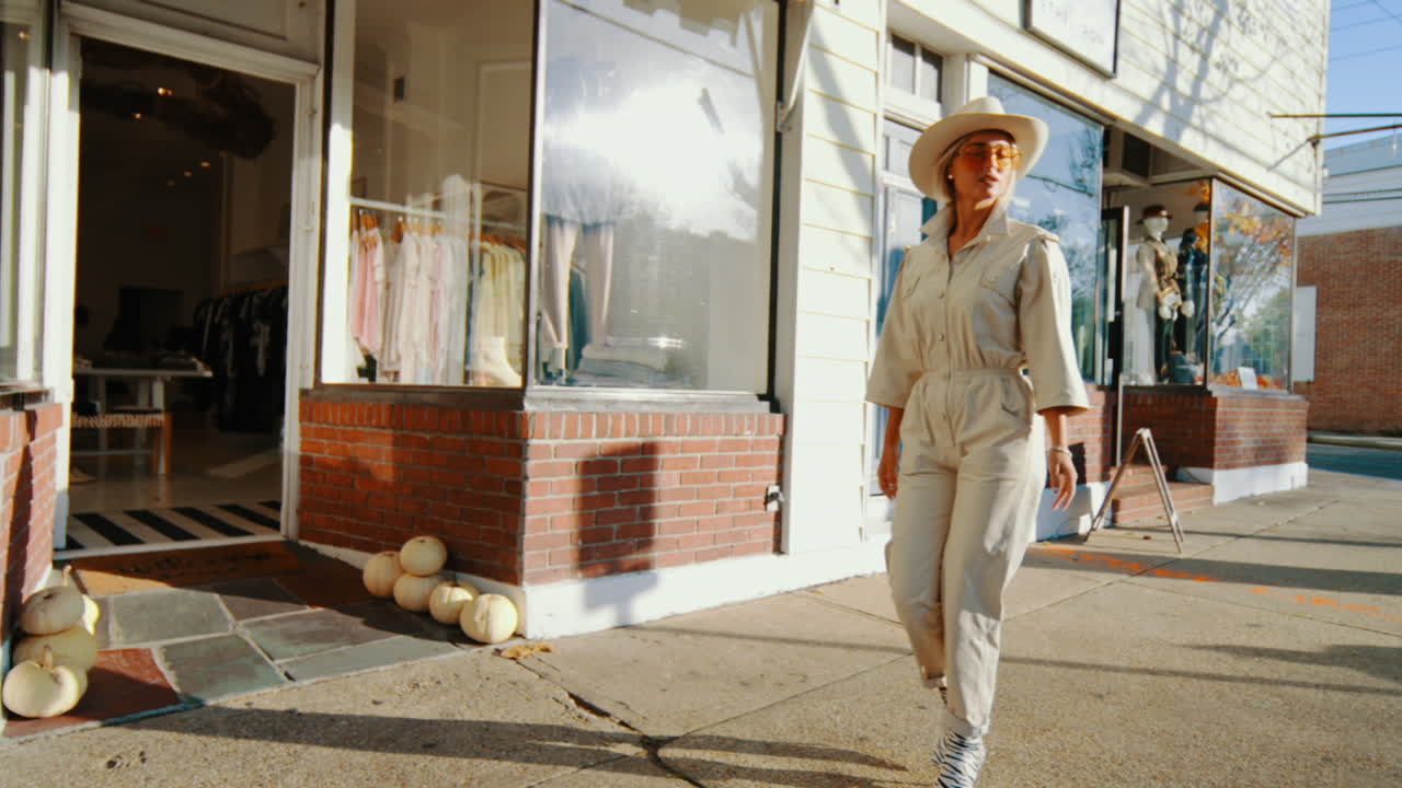 New York, USA, 2 maggio 2025: Donna alla moda che cammina davanti a una boutique con la porta blu. Inquadratura a figura intera di una donna in tuta beige e cappello da cowboy che cammina su un marciapiede illuminato dal sole davanti a negozi locali a New York