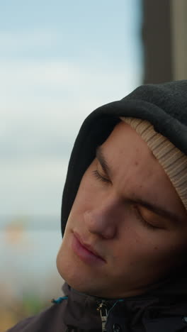 close-up of man turning head thoughtfully while wearing hoodie and beanie, with blurred background featuring modern office buildings