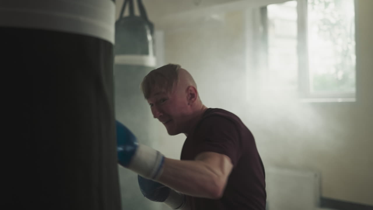 스포츠 센터에서 복싱 가방을 때리는 운동선수, 장갑을 입은 기쁜 운동선수는 연기가 은 체육관에서 팔로 매달린 장비를 때립니다. 느린 동작, 전투 공격 스타일