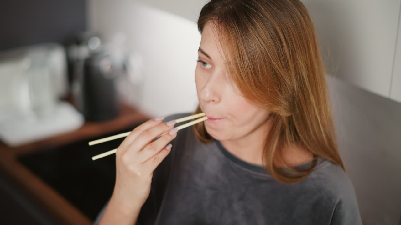 Hungry lady with long hair eating noodles using chopsticks while gazing attentively elsewhere in bright modern kitchen, kitchen appliances and utensils softly blurred in background