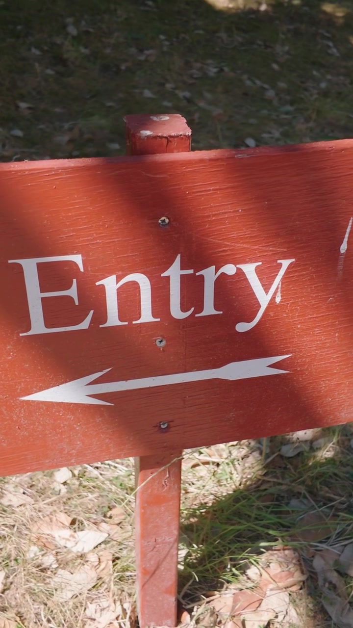 Señal de entrada con flecha izquierda en un tablero de madera naranja mientras la cámara se desplaza con una ligera órbita. Sombra moteada, camino de grava y césped sugieren señalización de entrada a un parque o recinto. Formato vertical.