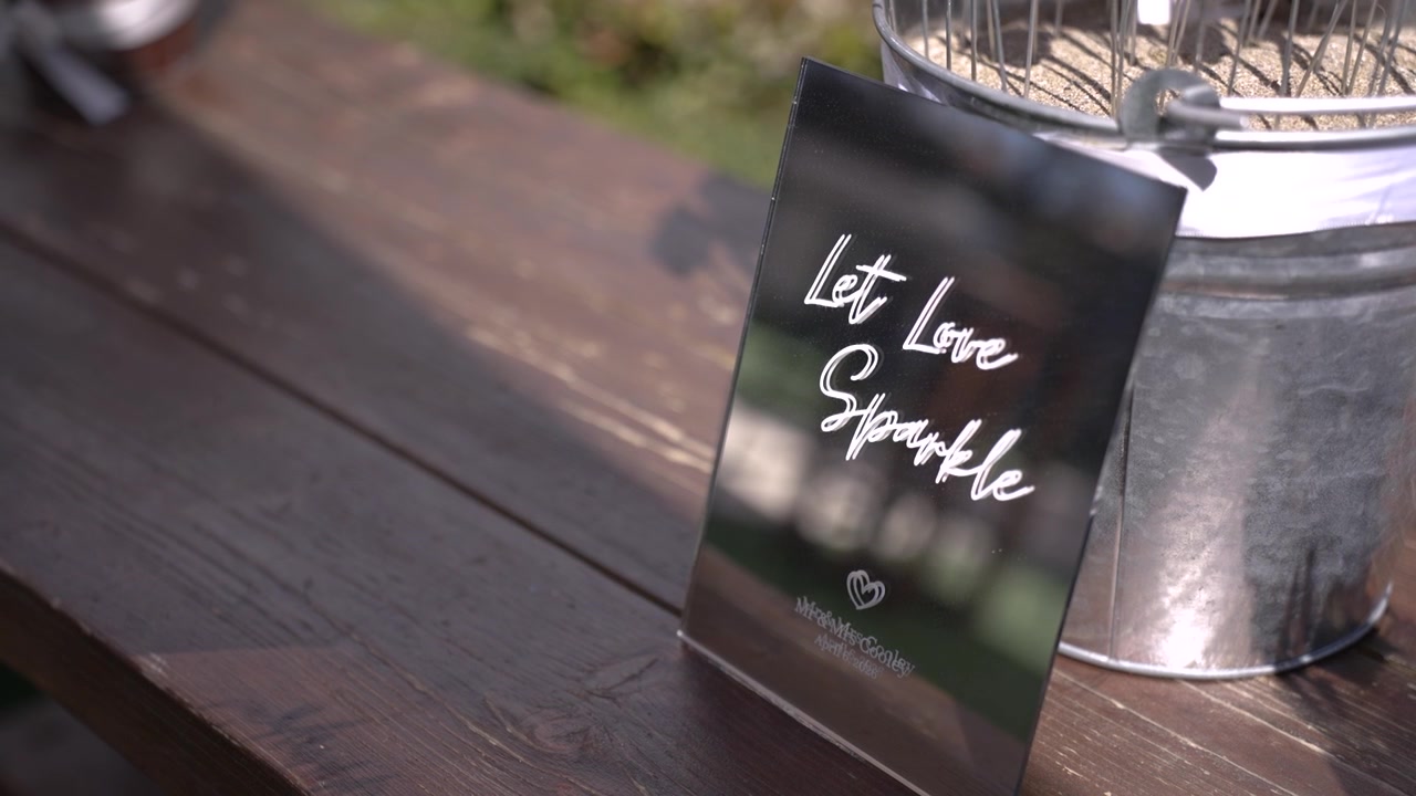 Et display viser et skilt, hvorpå der står "Lad kærligheden funkle", placeret på et bord. Omgivelserne er udendørs med grønt i baggrunden. Denne scene fanger et øjeblik ved en glædelig sammenkomst.