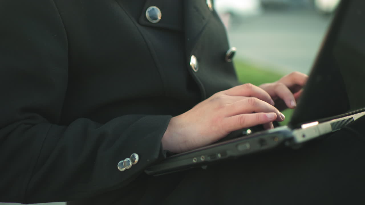 Close up of hands typing on laptop outdoors while wearing black suit with silver buttons, against blurred background featuring parked cars and greenery