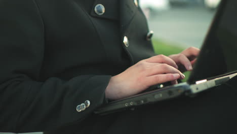 close up of hands typing on laptop outdoors while wearing black suit with silver buttons, against blurred background featuring parked cars and greenery