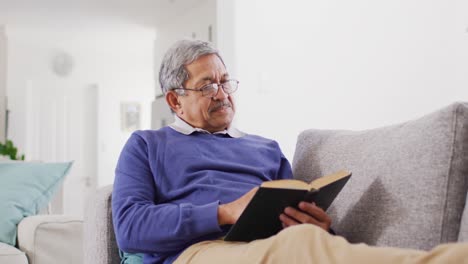 Video-De-Un-Feliz-Hombre-Birracial-Mayor-Relajándose-En-El-Sofá-Leyendo-Un-Libro-En-Casa