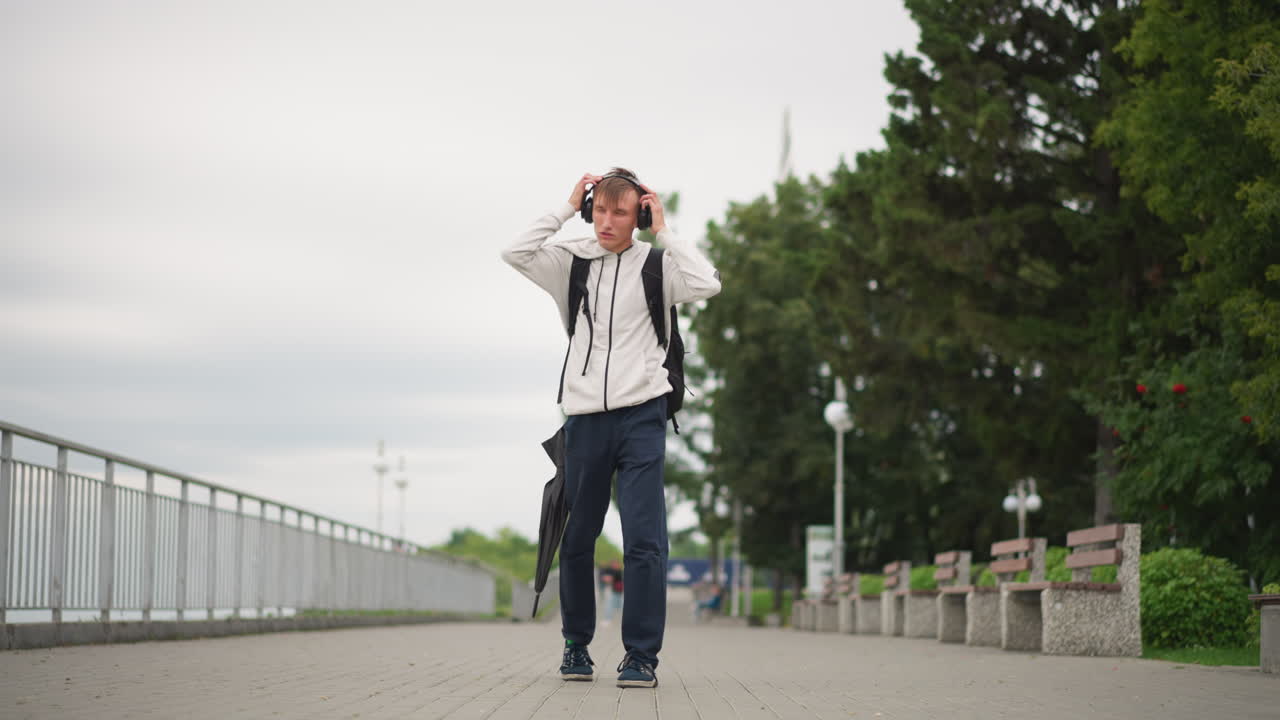 우산을 들고 산책로에서 잠시 멈춰선 백인 남성이 흐린 하늘 아래에서 수평선을 바라보며 헤드폰과 백팩을 정리하고 있어요. 프레임에는 통근자가 스케줄을 확인하고, 콘텐츠 제작자가 장비를 테스트하는 모습이 담겨 있습니다.