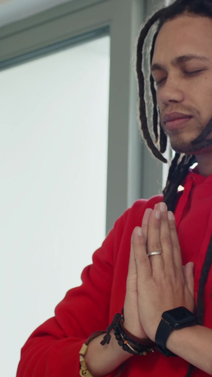 Vertical video: Soft light drawing man meditating near frosted pane in red hoodie, watch