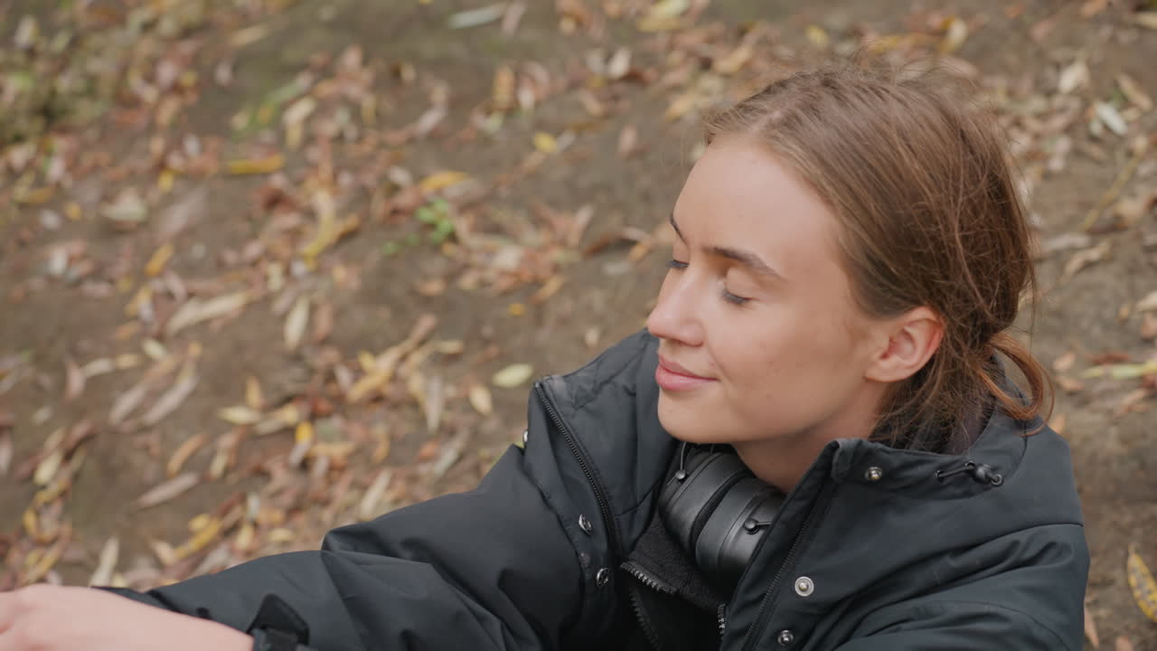 outdoor autumn scene caucasian woman sitting among fallen leaves wearing headphones and dark jacket, looking upward with thoughtful expression, soft overcast light, earthy tones, student taking