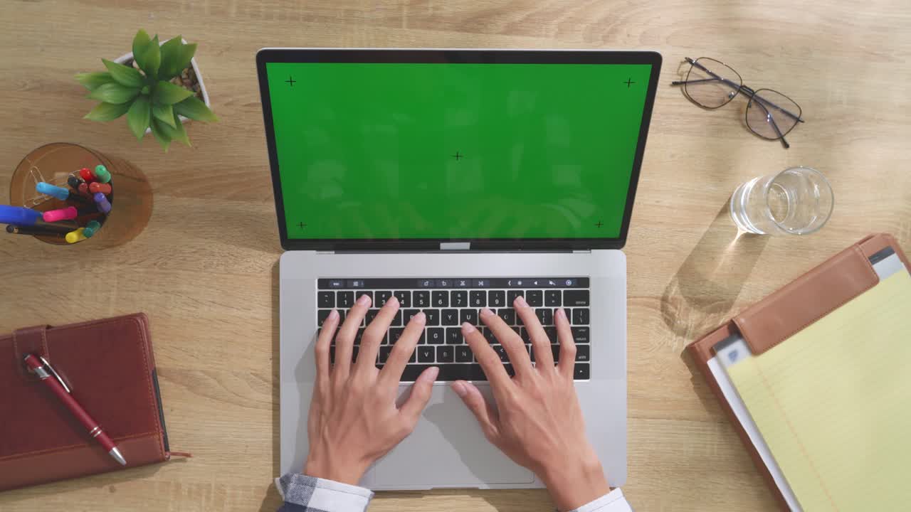 vista de arriba hacia abajo de una computadora portátil con pantalla verde simulada pantalla chromakey en un escritorio de oficina de madera al lado de la libreta con bolígrafos, gafas y un vaso de agua.