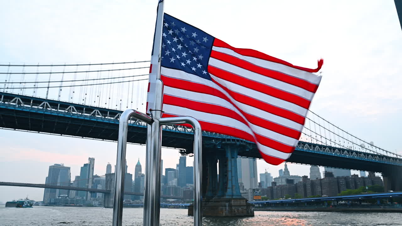 Flag waving by Brooklyn Bridge. Waves of an American flag are visible as a boat passes under the Brooklyn Bridge at dusk with a city skyline backdrop
