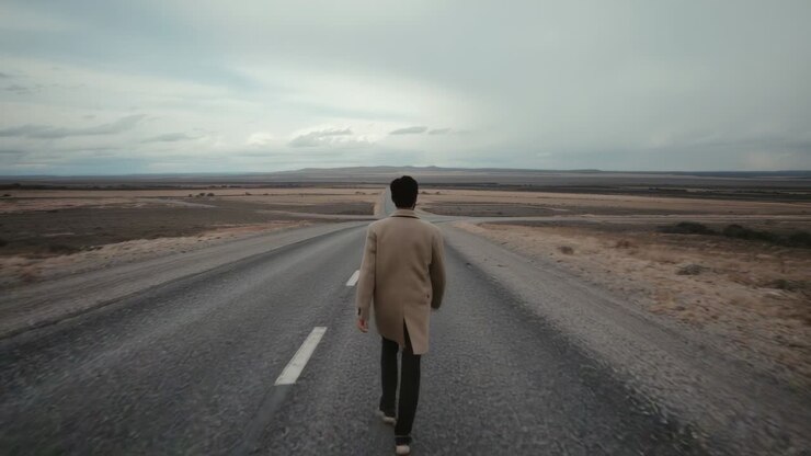 Camera starting, walking man in beige coat heading toward horizon on two-lane road, with rain shaft