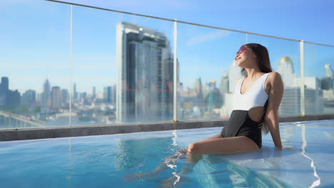 beautiful asian woman sitting in rooftop swimming pool, city skyline